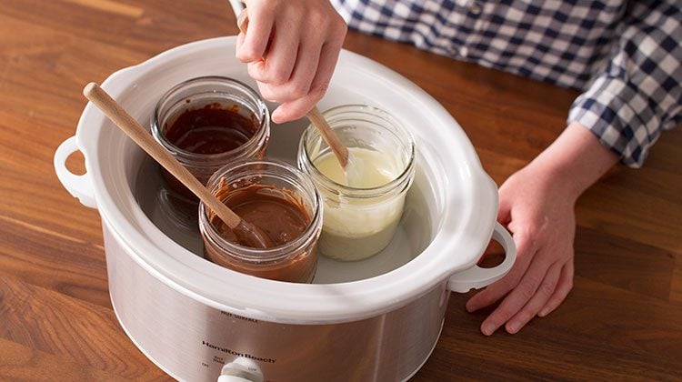 Slow cooker with three glass jars of different types of melted chocolate inside them including melted milk, melted white and melted dark
