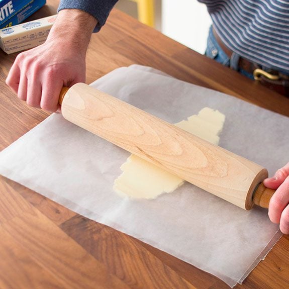 person using a rolling pin to flatten a stick of butter between two pieces of waxed paper