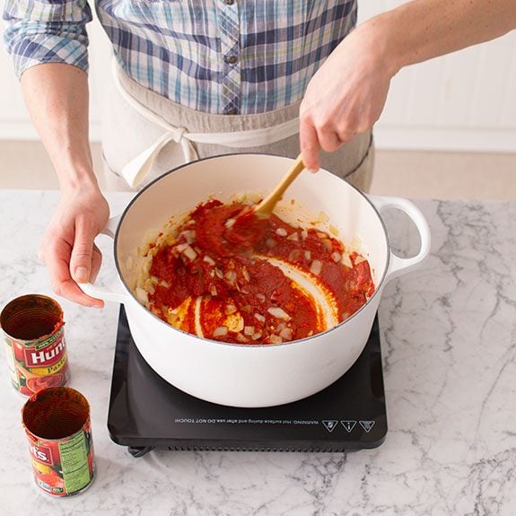 Tomato paste being stirred with a wooden spoon into a mixture of onions and garlic inside a dutch oven