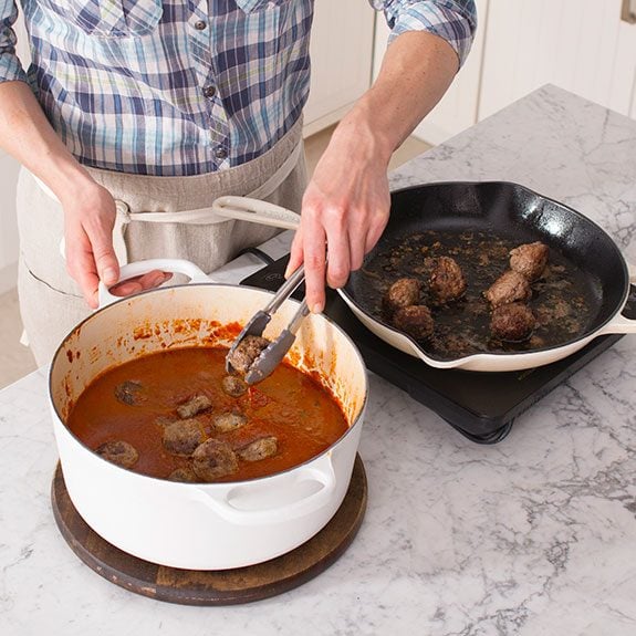 Using tongs, the person then transfers their meatballs into the dutch oven filled with their sauce to submerge them