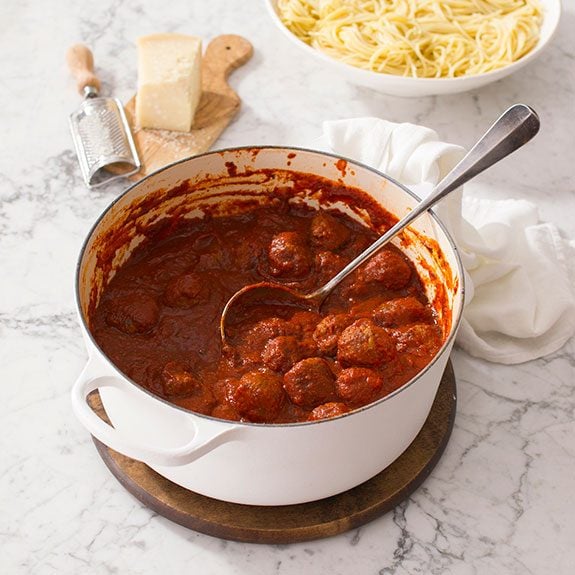 Dutch oven filled with sauce-covered meatballs and a large metal spoon resting inside for serving. Plain pasta waits in a bowl behind