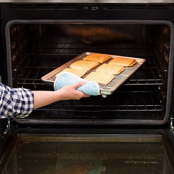 Waffles lined up on a wire rack on a baking sheet about to be set down inside an oven